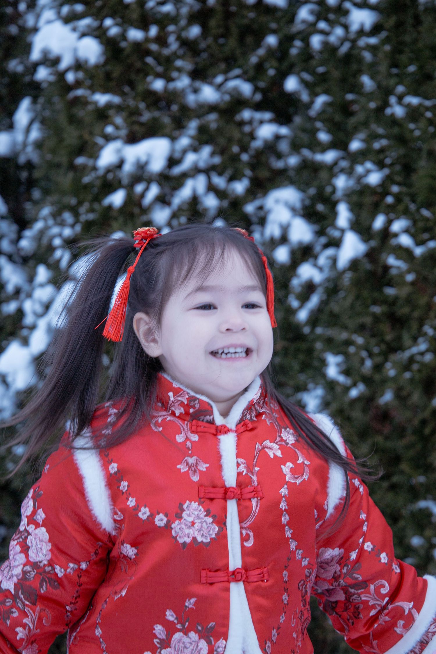 Young child in red traditional Chinese dress smiling in snowy winter scene.