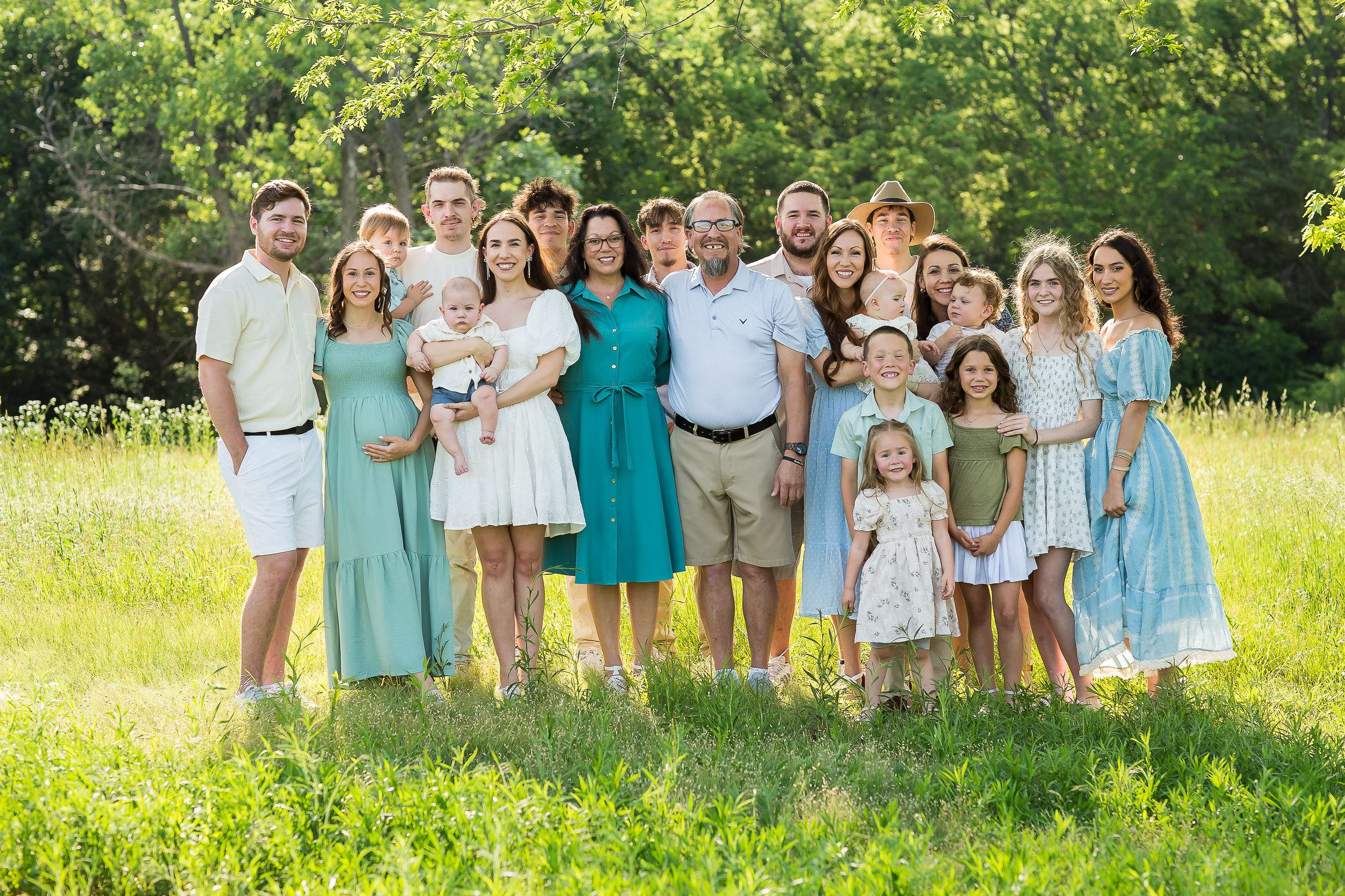 Large family group portrait outdoors in green meadow, wearing coordinated teal and white outfits, smiling together.
