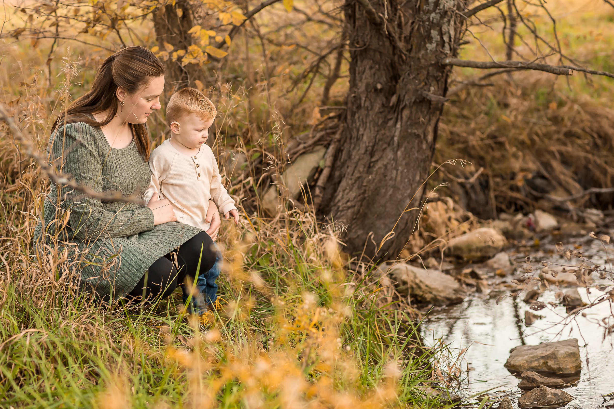 Mother and young child sitting together by a peaceful stream near a tree in an autumn outdoor setting.