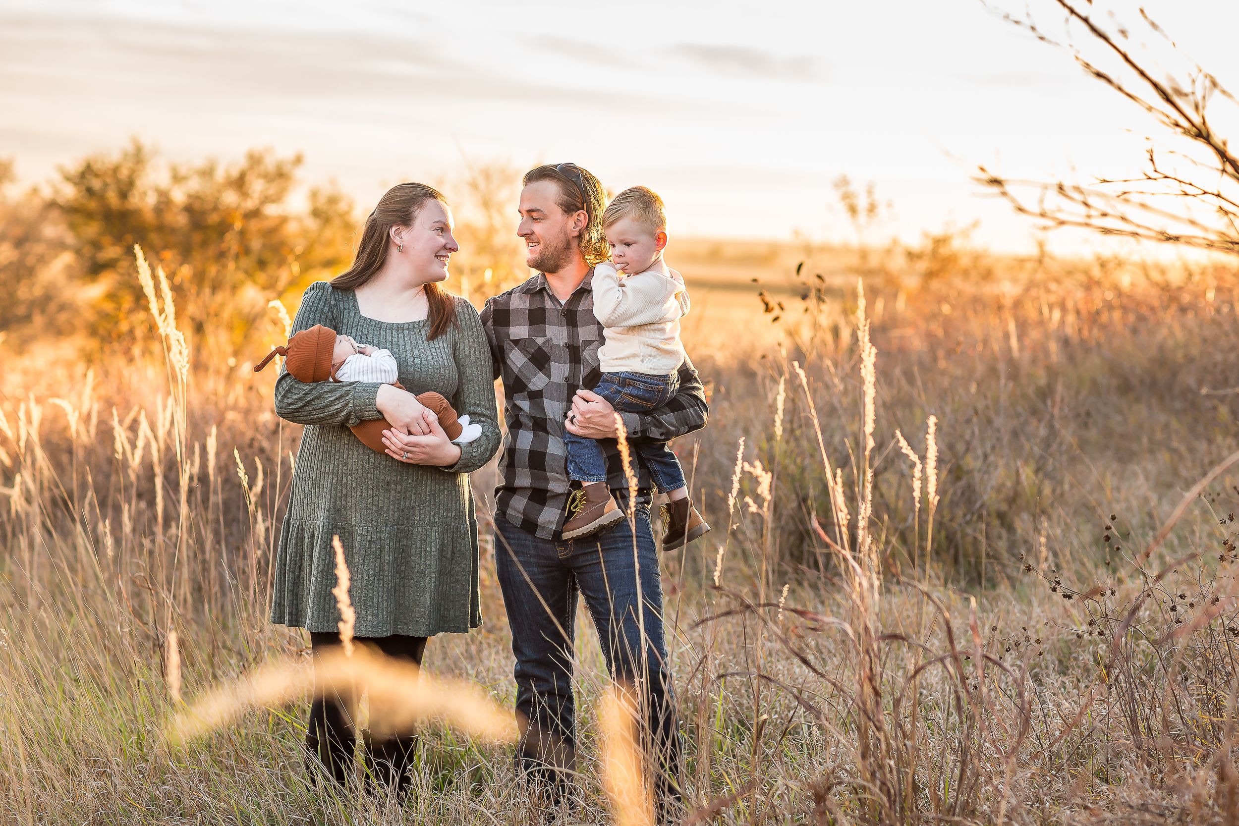 Family of four poses in golden field at sunset, parents holding two young children, smiling warmly.