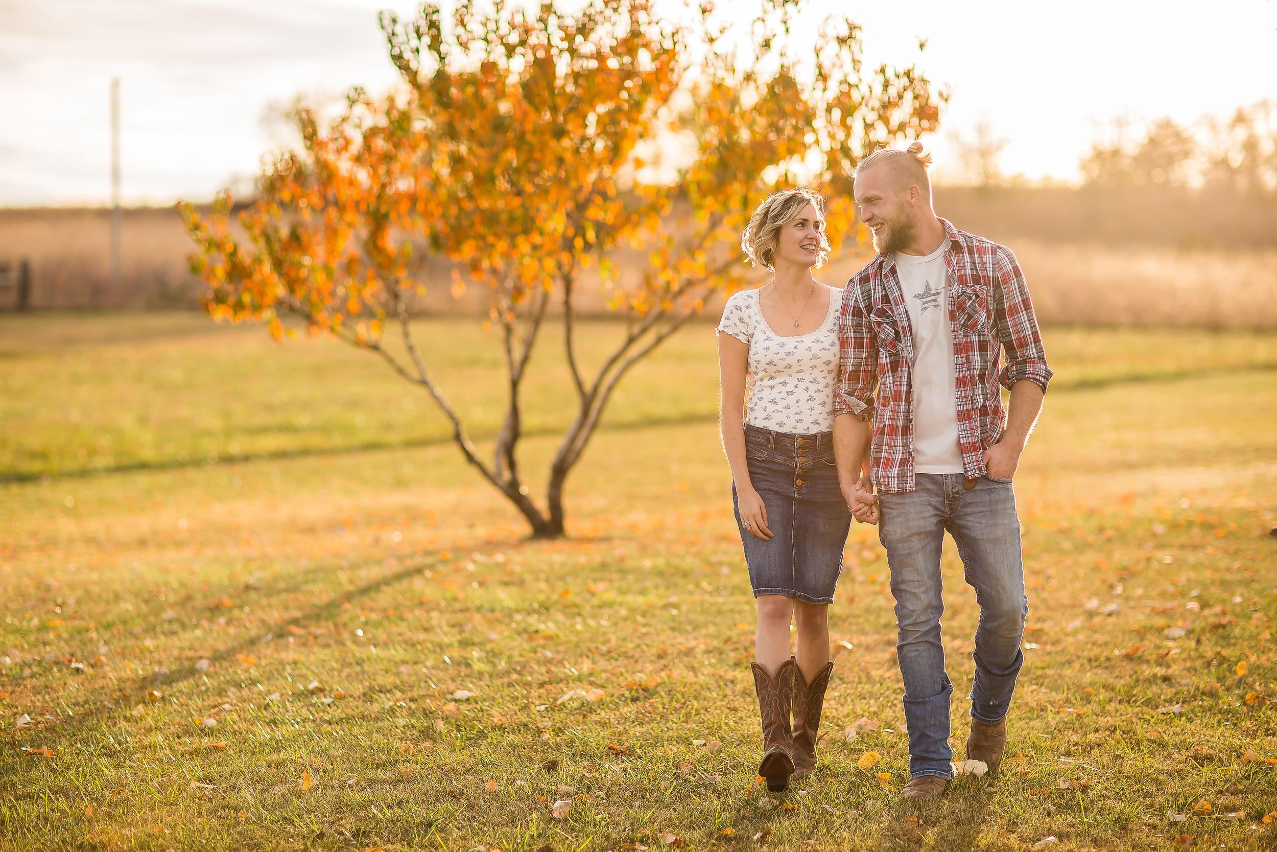 Couple walking hand-in-hand in a golden autumn field with a colorful tree and warm sunset light.