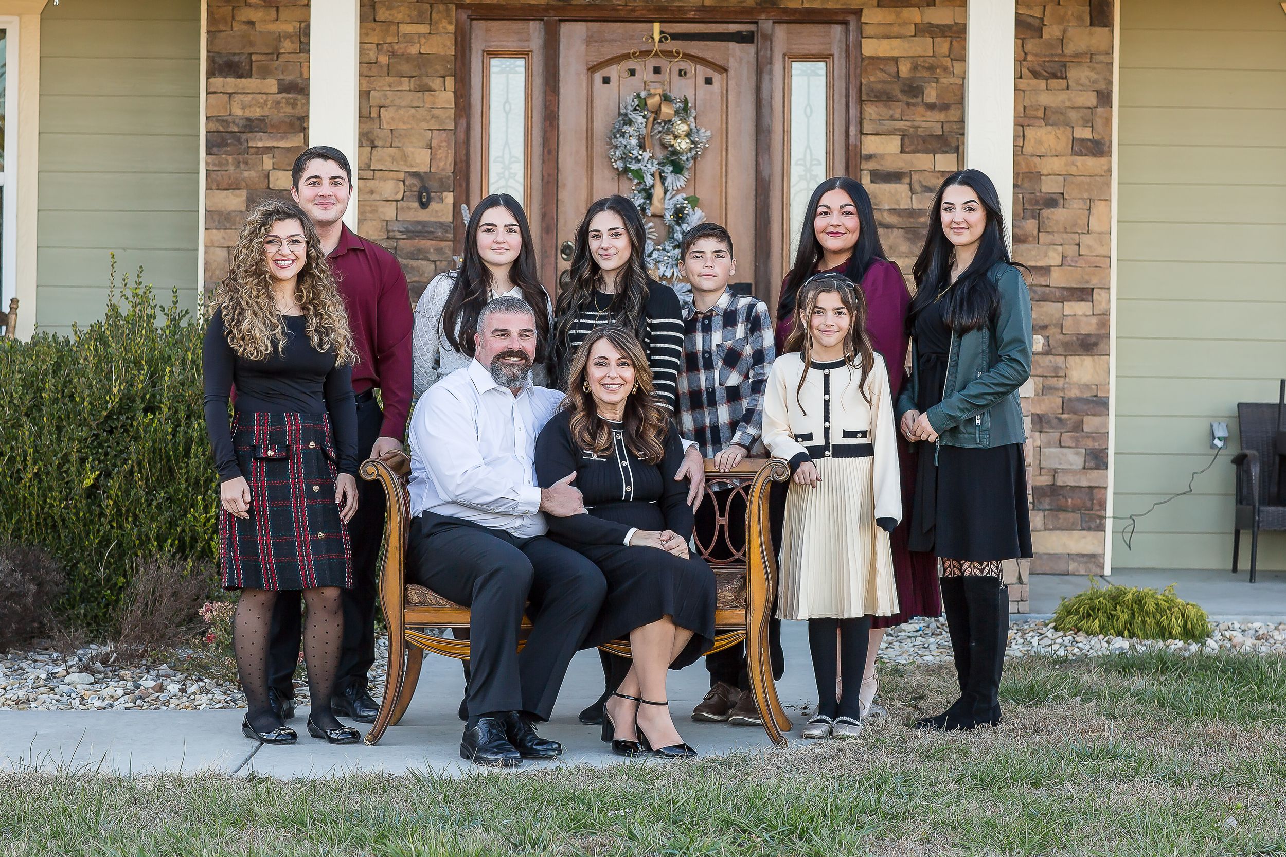 Large family portrait of parents and seven children posing together in front of a brick home with holiday decor.