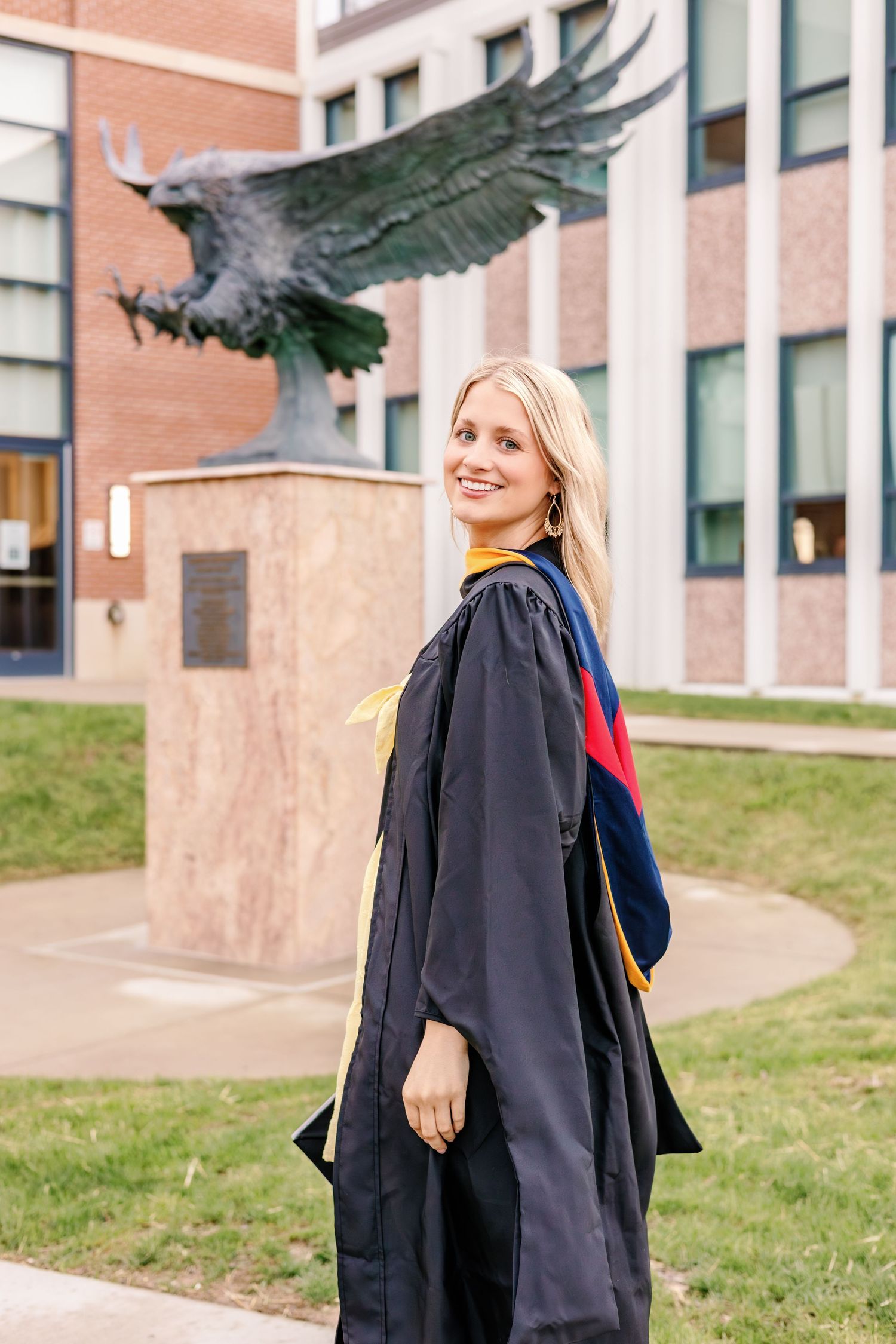 A graduate in a black gown poses in front of a brick building on campus.
