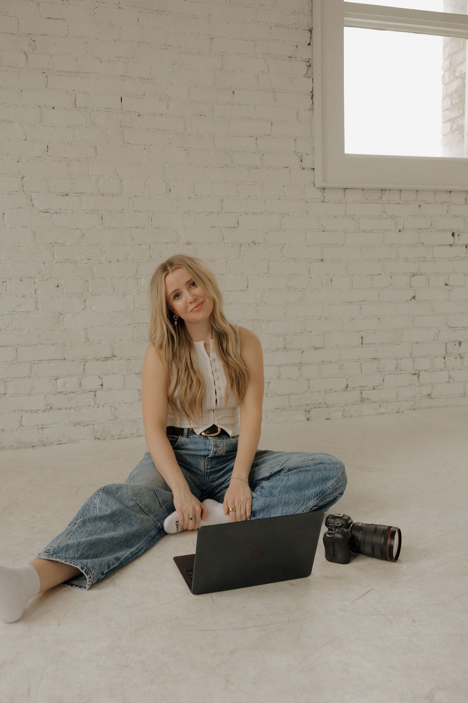 Blonde female photographer sitting cross-legged on floor with laptop and camera in white brick studio.