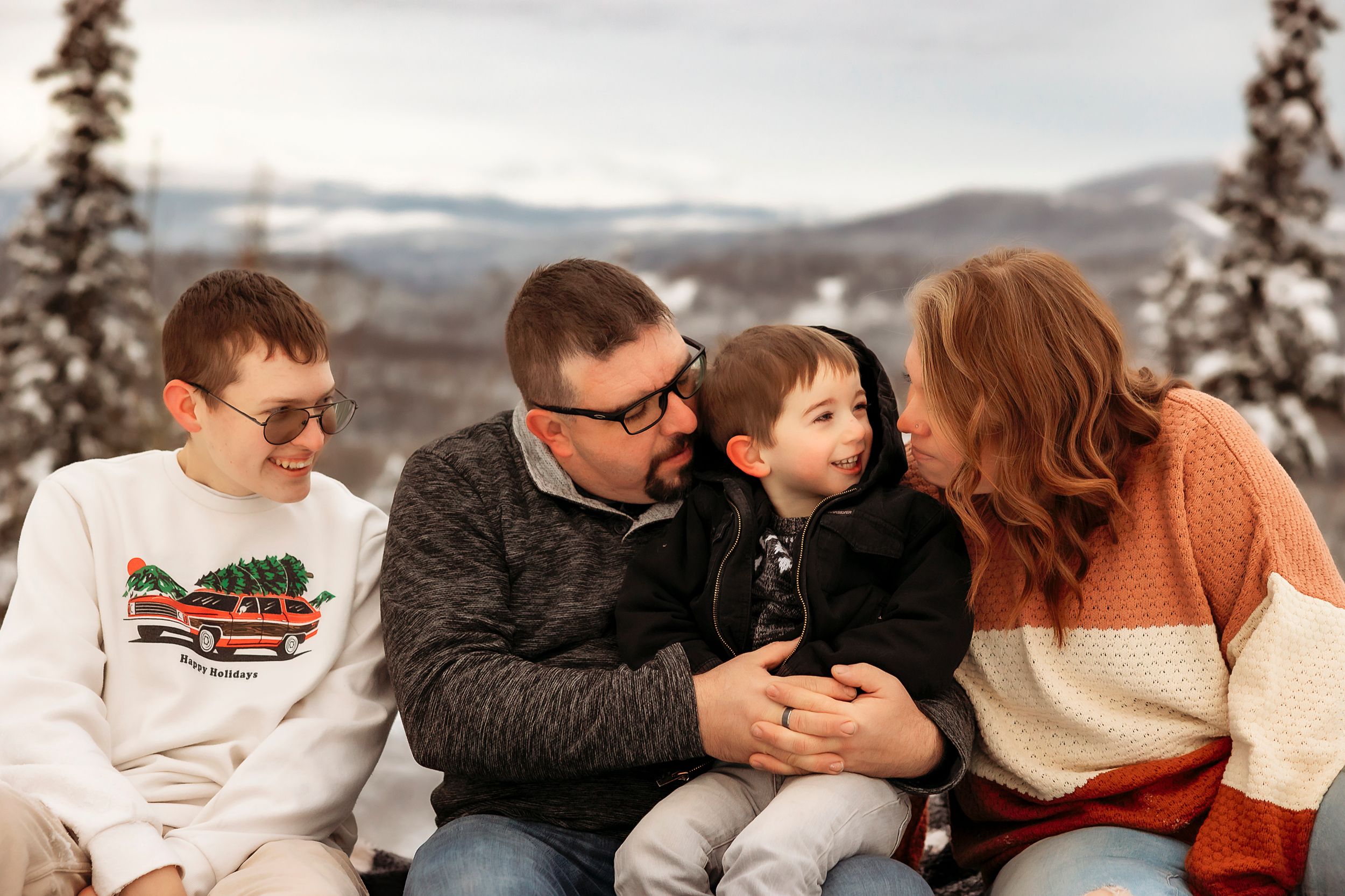 A winter family portrait session with people sitting close together in warm clothing against a snowy mountain backdrop.