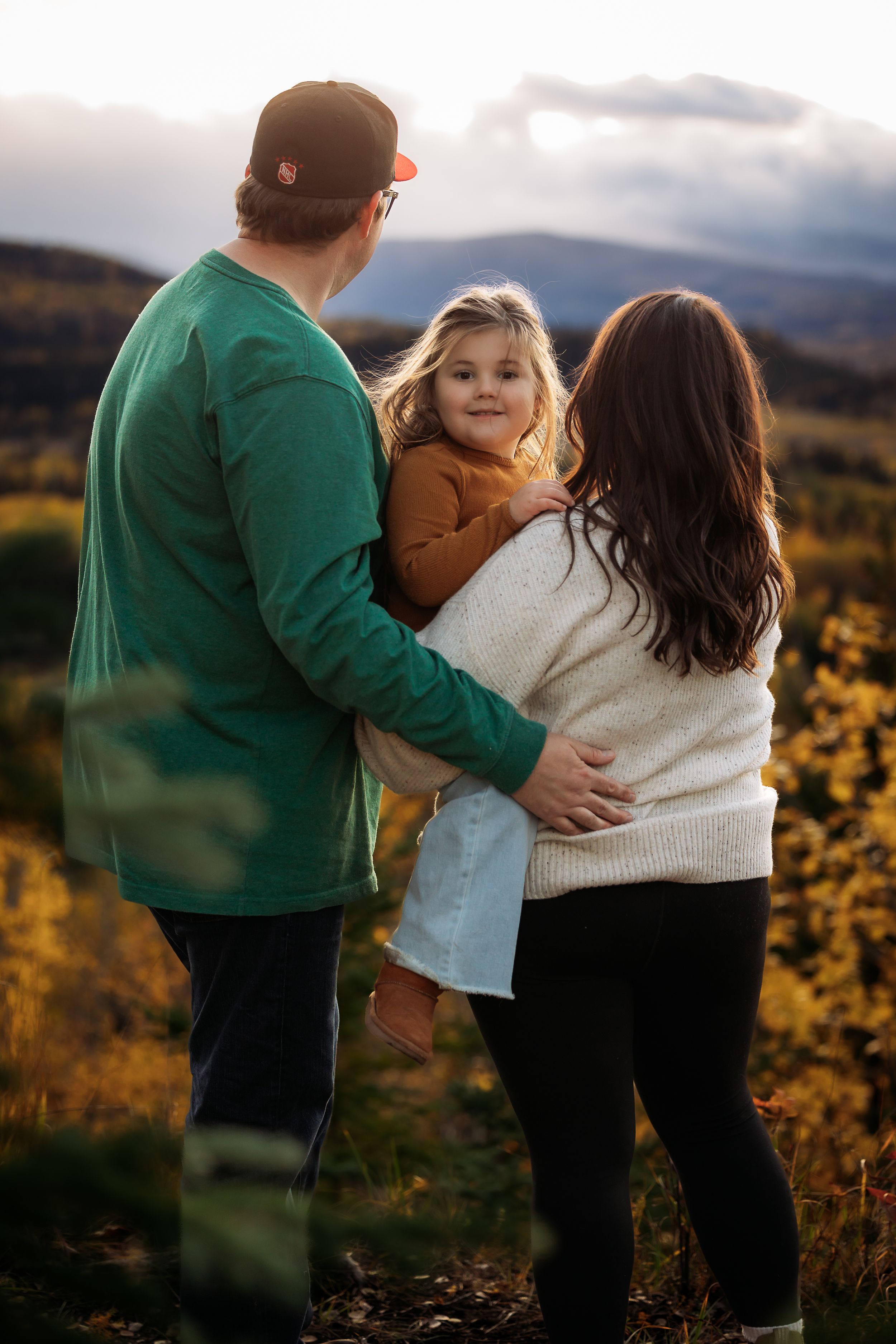 A family embraces while standing on a hilltop during golden hour with autumn foliage in the background.