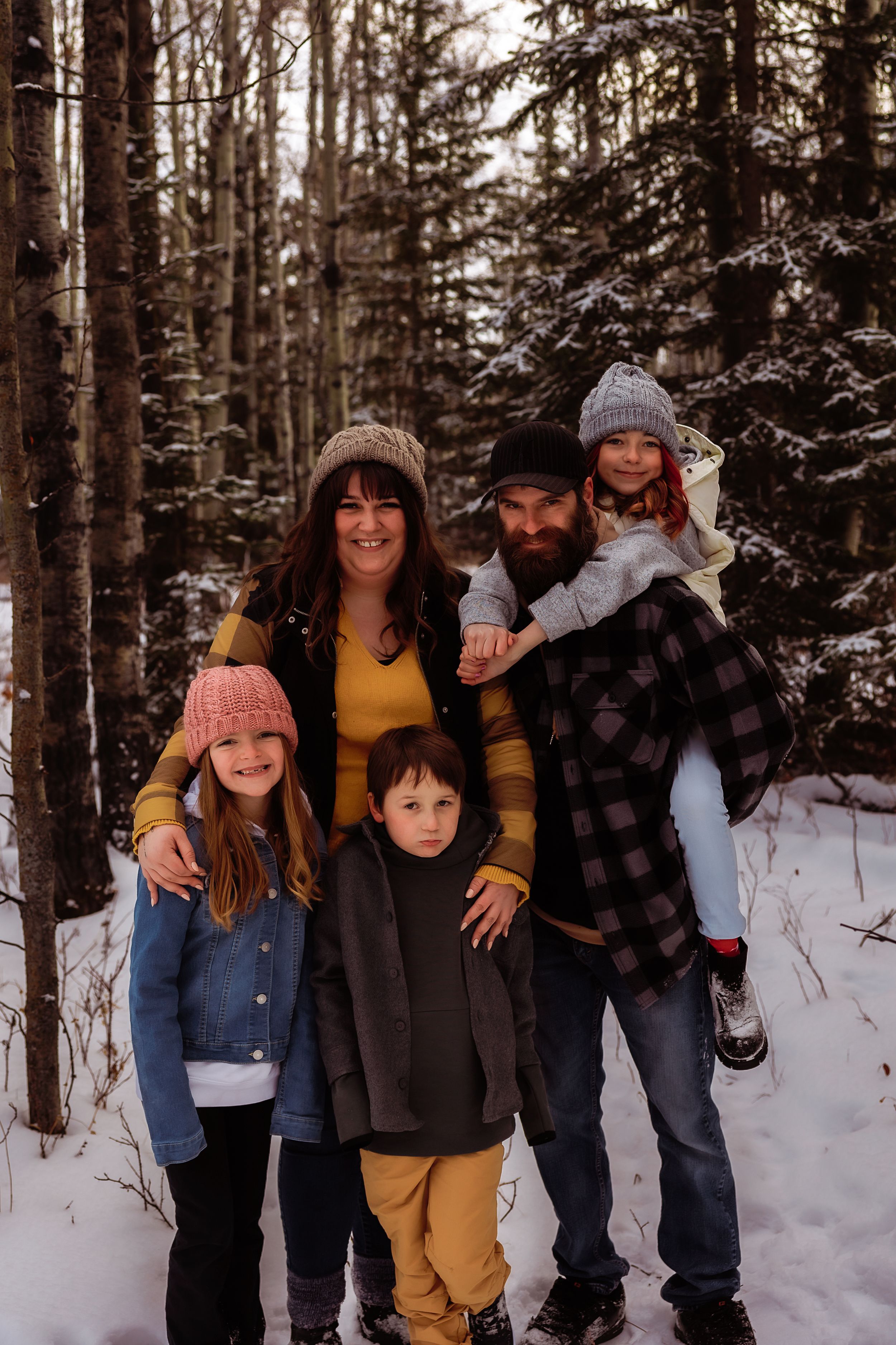 A family poses together in a snowy winter forest setting wearing warm clothing and knit hats.