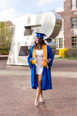 Graduate in blue cap and gown poses in front of large sculptural letter B on brick plaza during commencement.