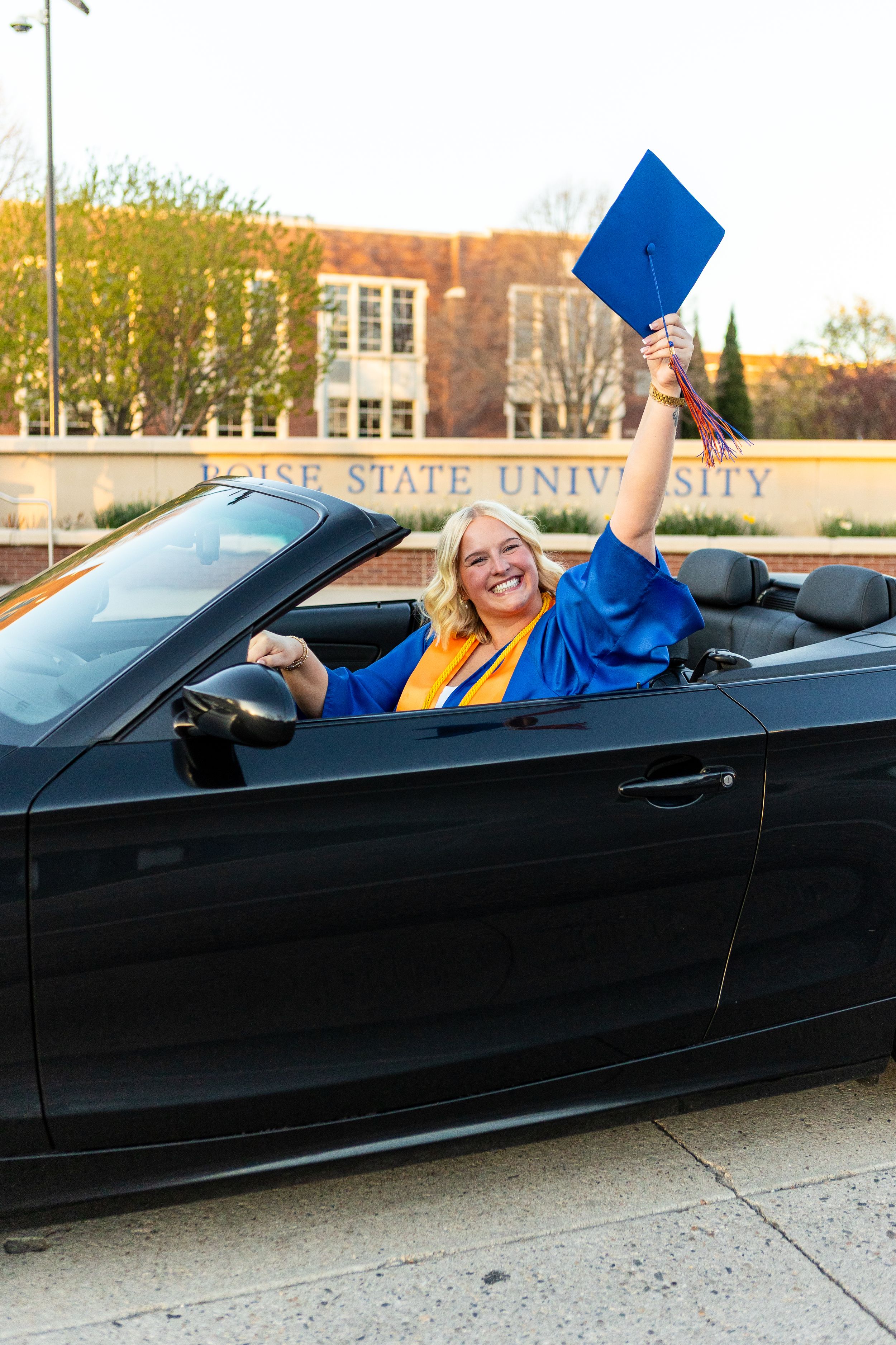 Graduate in blue and gold regalia celebrates in black convertible car outside university building.