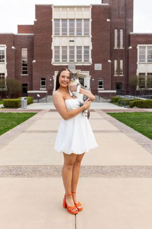 Someone in a white dress holding a diploma stands outside a brick school building on graduation day.