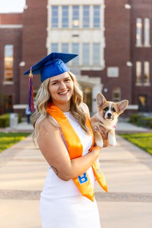 Graduate in white dress and blue cap holds small corgi while posing on campus with orange graduation sash.