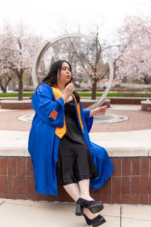 A graduate wearing blue academic regalia sits on brick wall near blooming cherry blossom trees on a spring day.