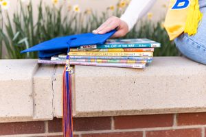 A stack of colorful books and school supplies resting on a brick ledge with foliage in the background.
