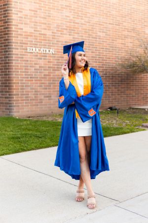 Graduate wearing a bright blue cap and gown with gold stole poses confidently on a campus sidewalk.