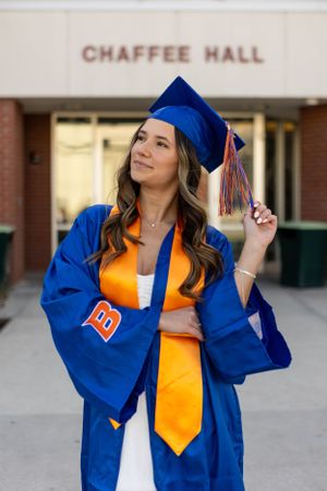 Graduate in royal blue cap and gown with orange sash poses outside Chaffee Hall during commencement ceremony.