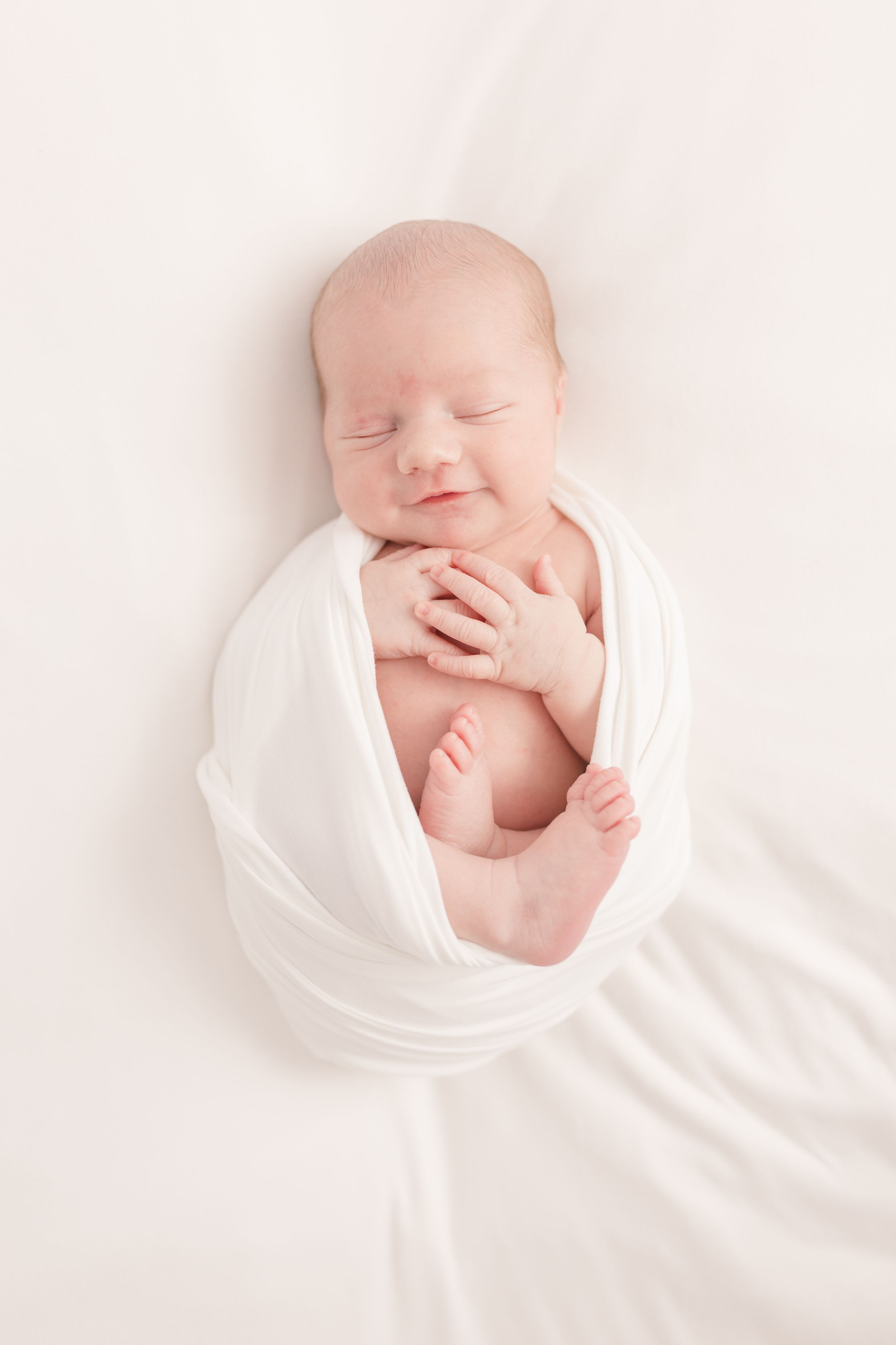 Peaceful newborn baby wrapped in soft white fabric against cream background.