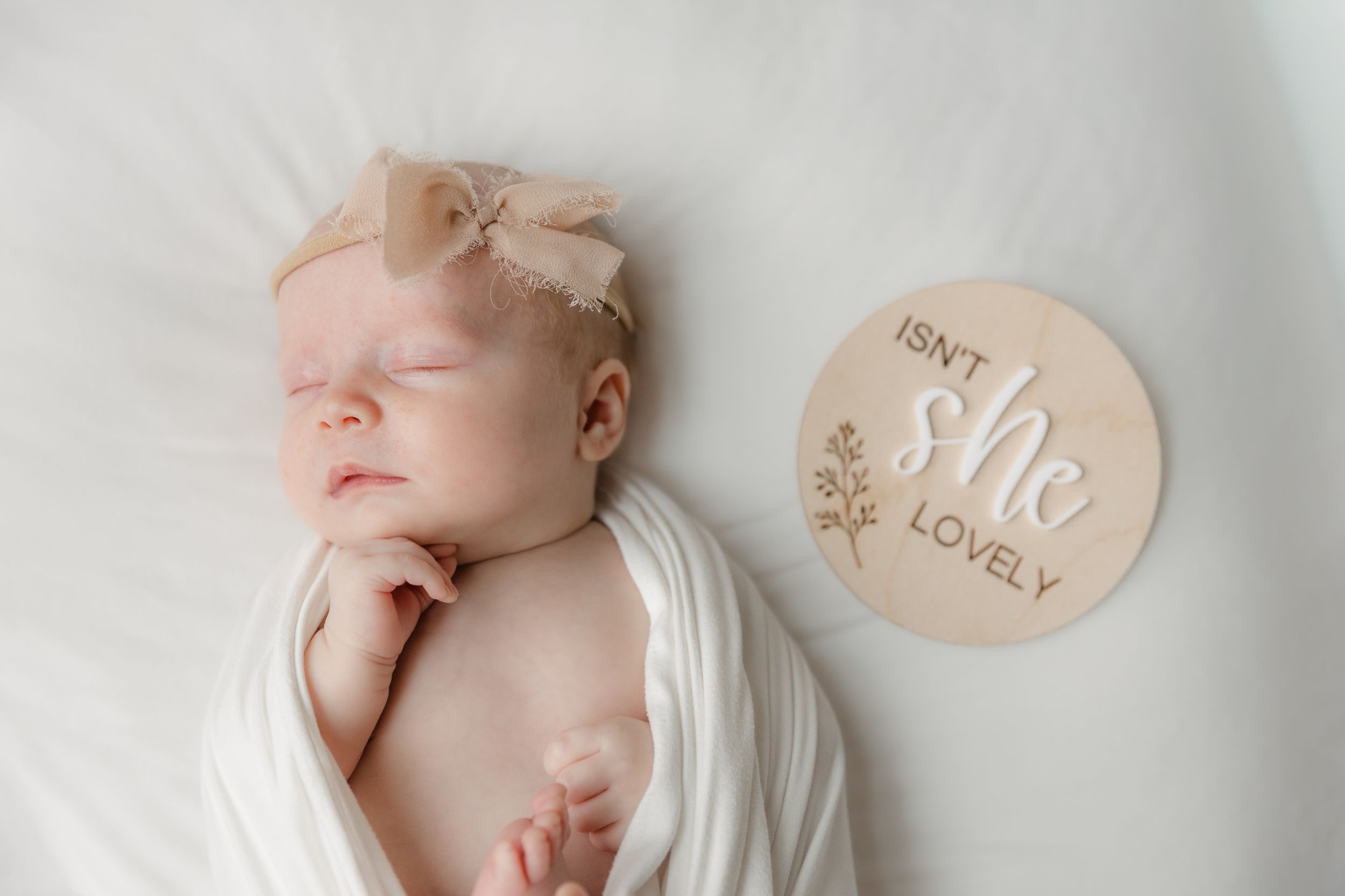 Newborn baby girl swaddled in white wrap with bow headband beside a wooden milestone disc reading Isn't She Lovely.