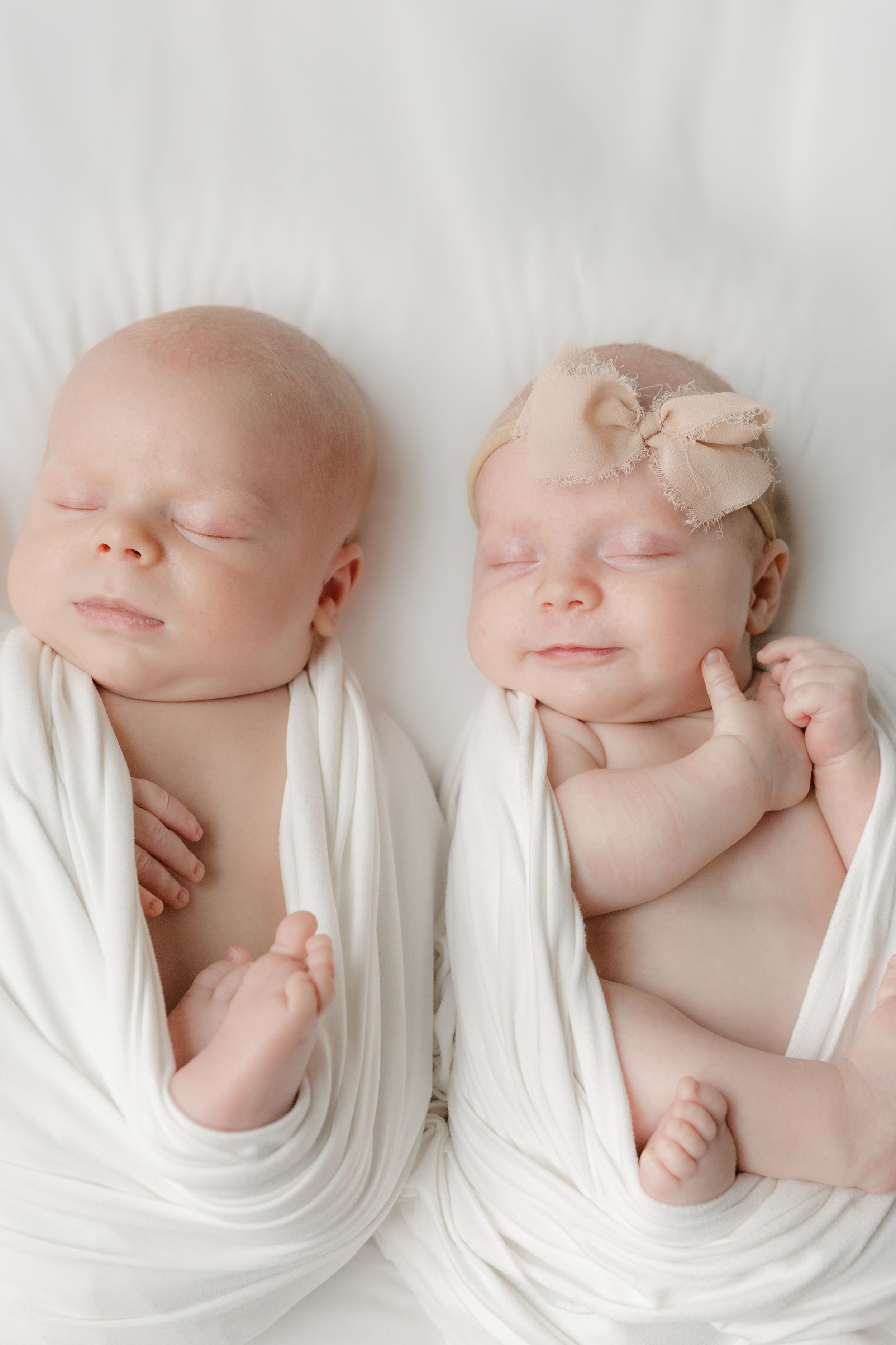 Two sleeping newborn twins swaddled in white wraps, one wearing a bow headband, lying side by side on a white surface.