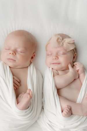 Two sleeping newborn twins swaddled in white wraps, one wearing a bow headband, lying side by side on a white surface.