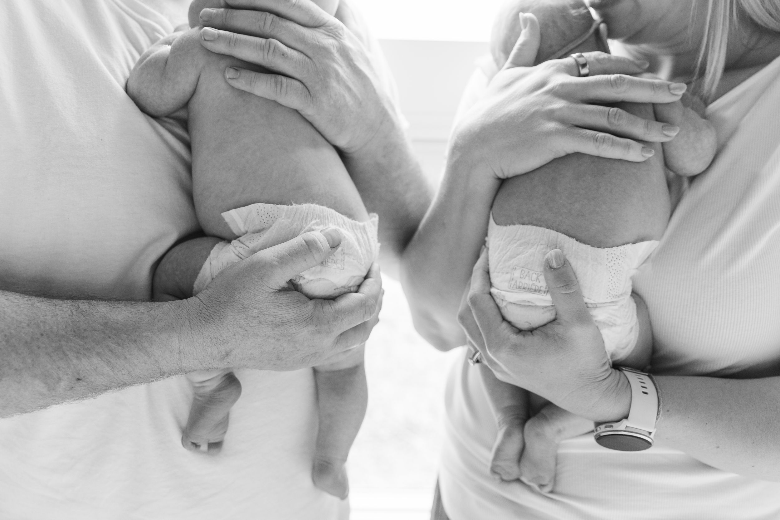 Black and white photo of two adults each holding a newborn baby, showing tiny feet and hospital bracelets.