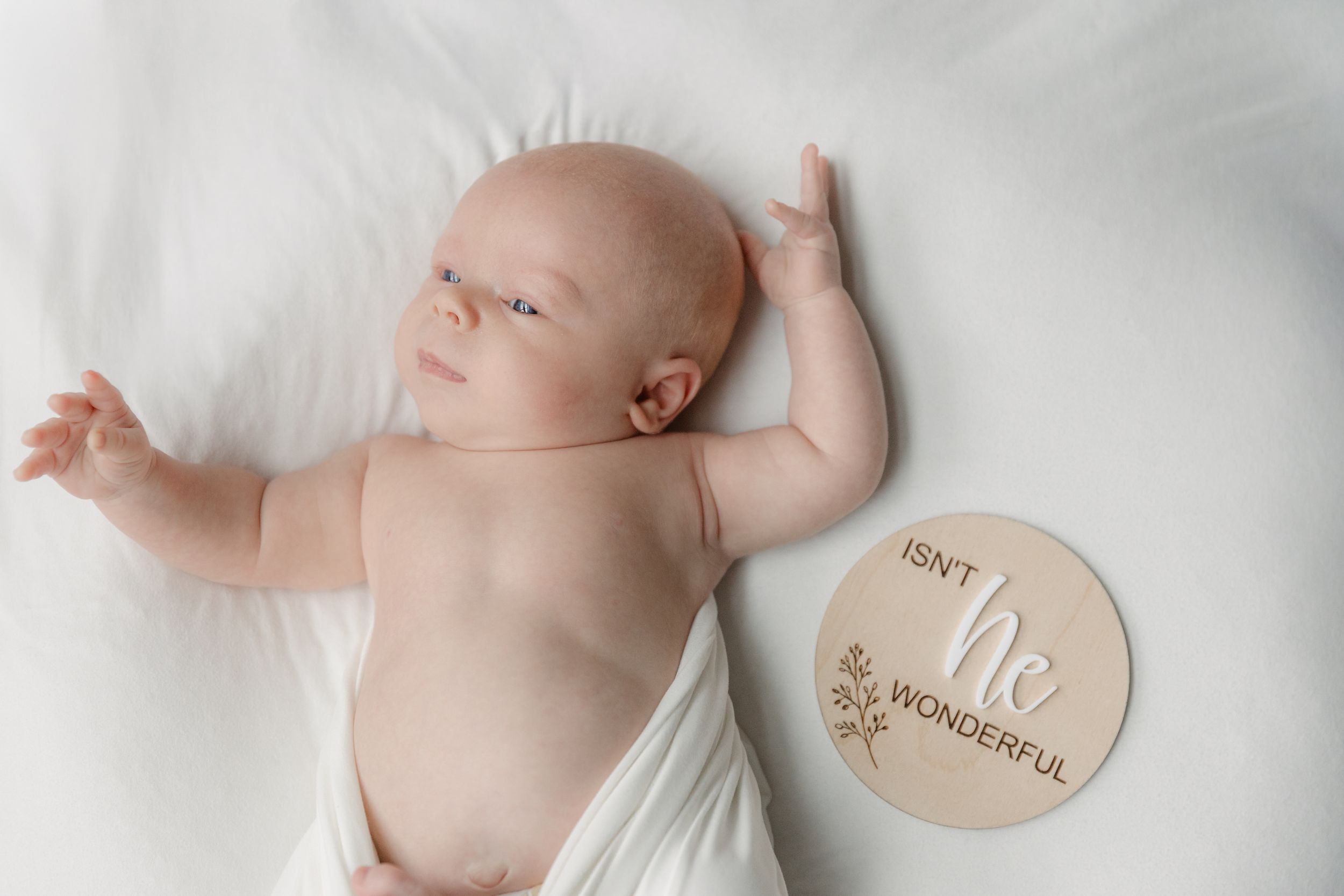 Newborn baby boy lying on white blanket beside a wooden round sign reading Isn't He Wonderful.