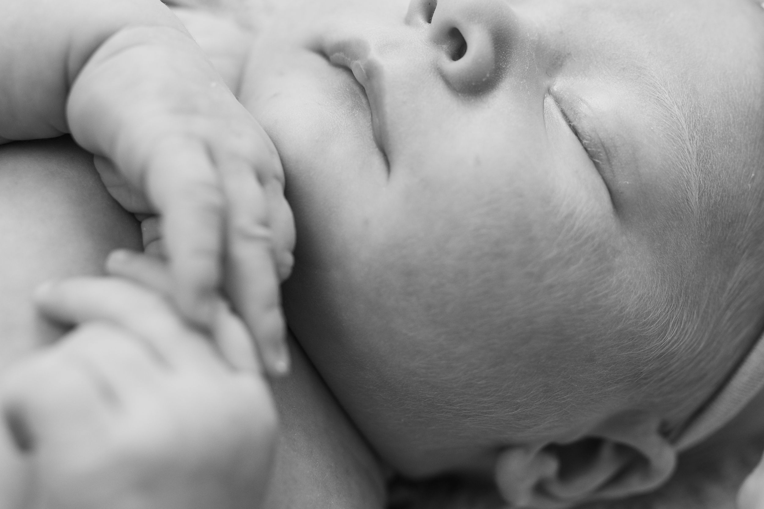 Close-up black and white photo of a sleeping newborn baby with tiny hands tucked near their face.