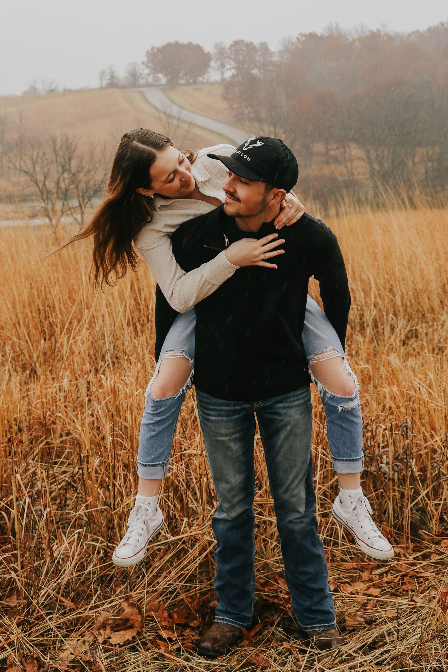 Playful piggyback moment between couple in autumn field with rolling hills in background.