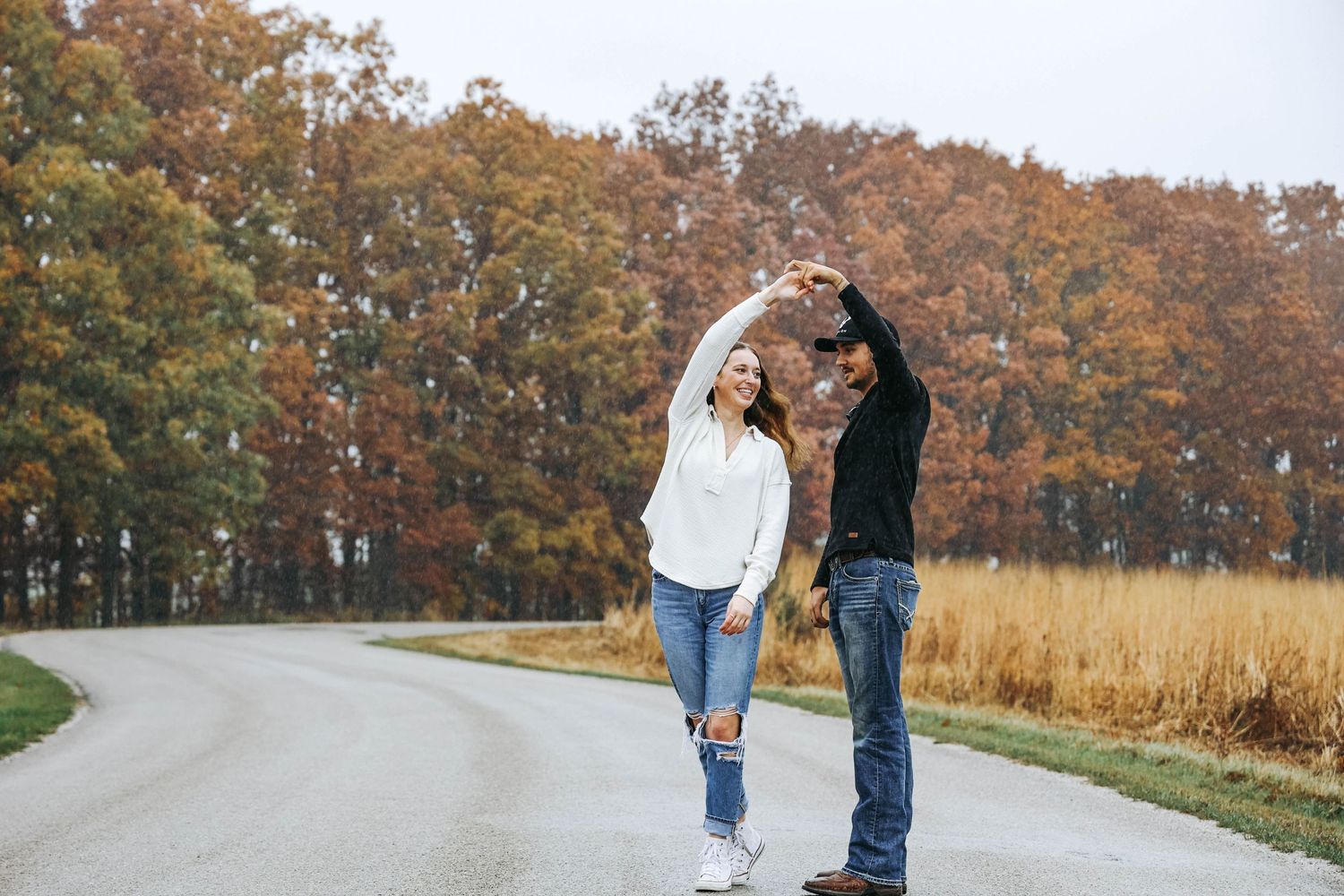 A romantic couple dances together on a winding country road surrounded by autumn trees.