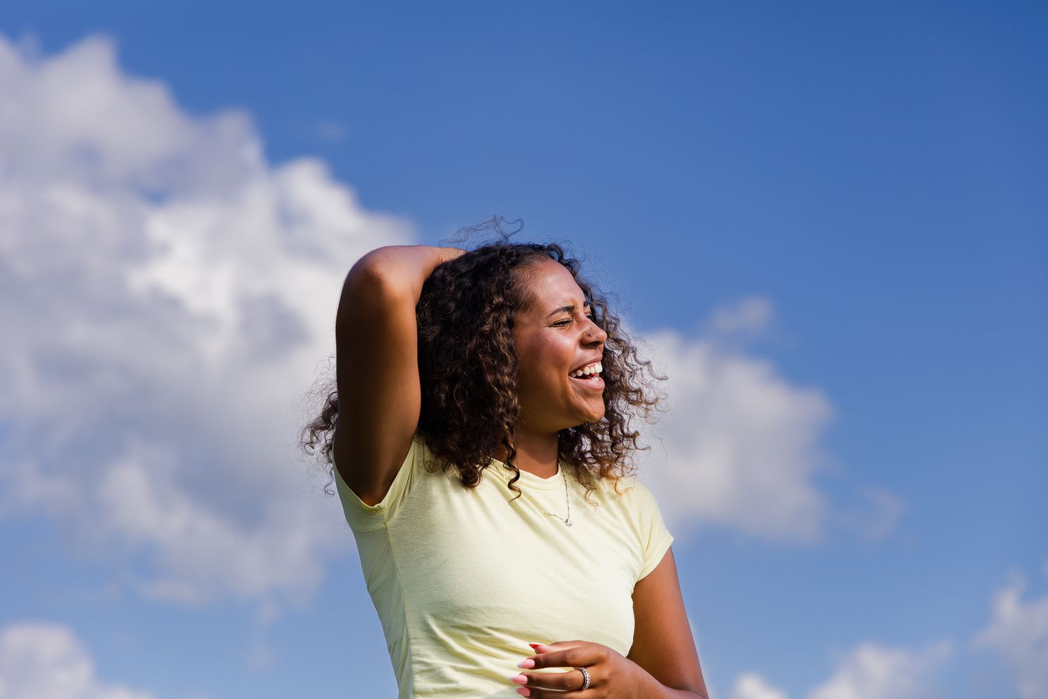 Woman in yellow shirt laughing with joy against bright blue sky with white puffy clouds.