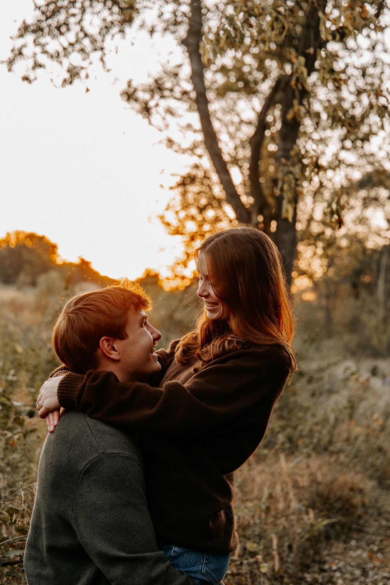 A couple embraces in a warm sunset glow among autumn trees and tall grass in a romantic outdoor setting.