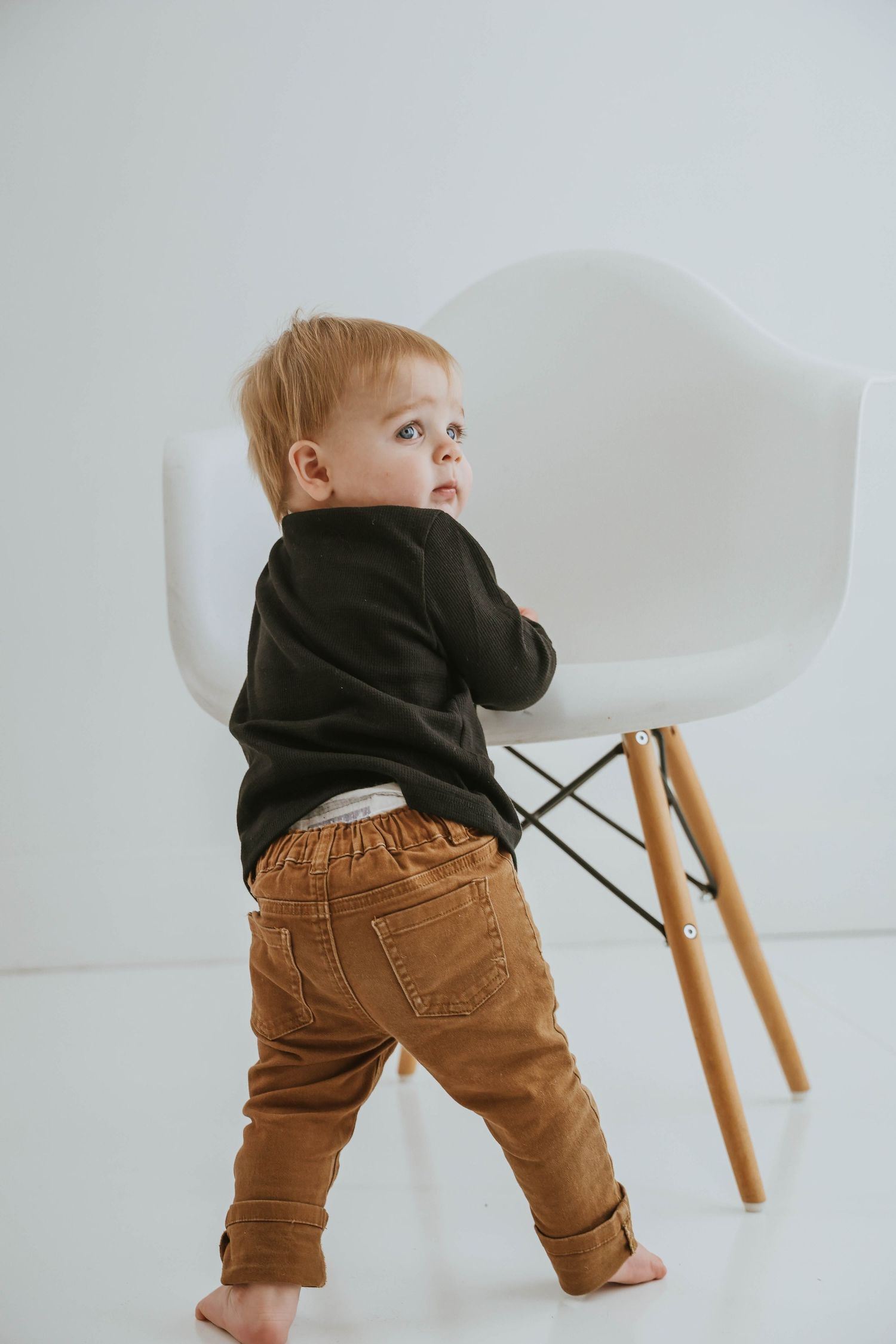 Toddler in black sweater and brown pants standing by modern white chair.