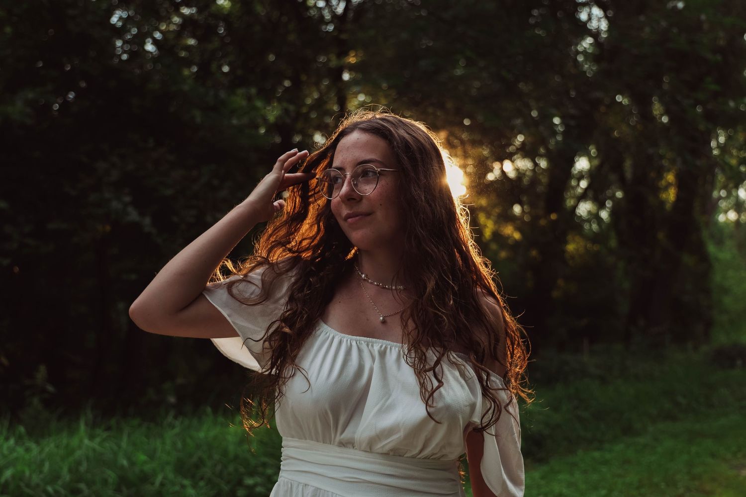 Woman in white dress brushing hair from face in golden sunset light.
