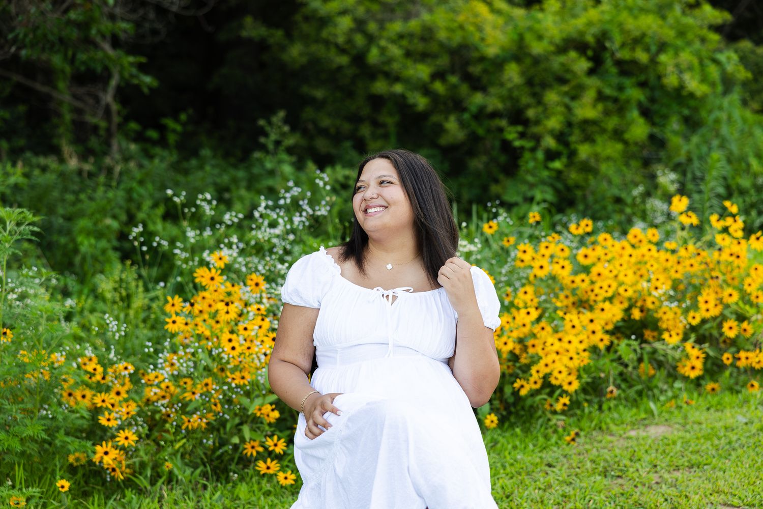 Person in a white dress poses gracefully in a garden filled with vibrant yellow flowers on a sunny day.