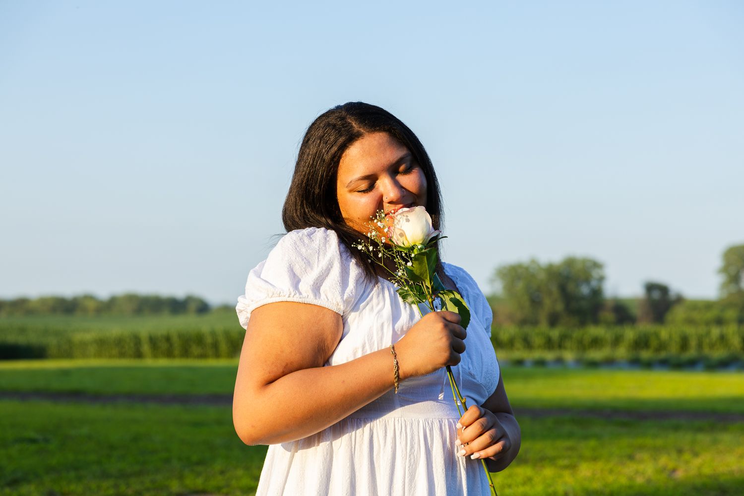 A person in a white dress smelling a bouquet of flowers in a sunny field during golden hour.