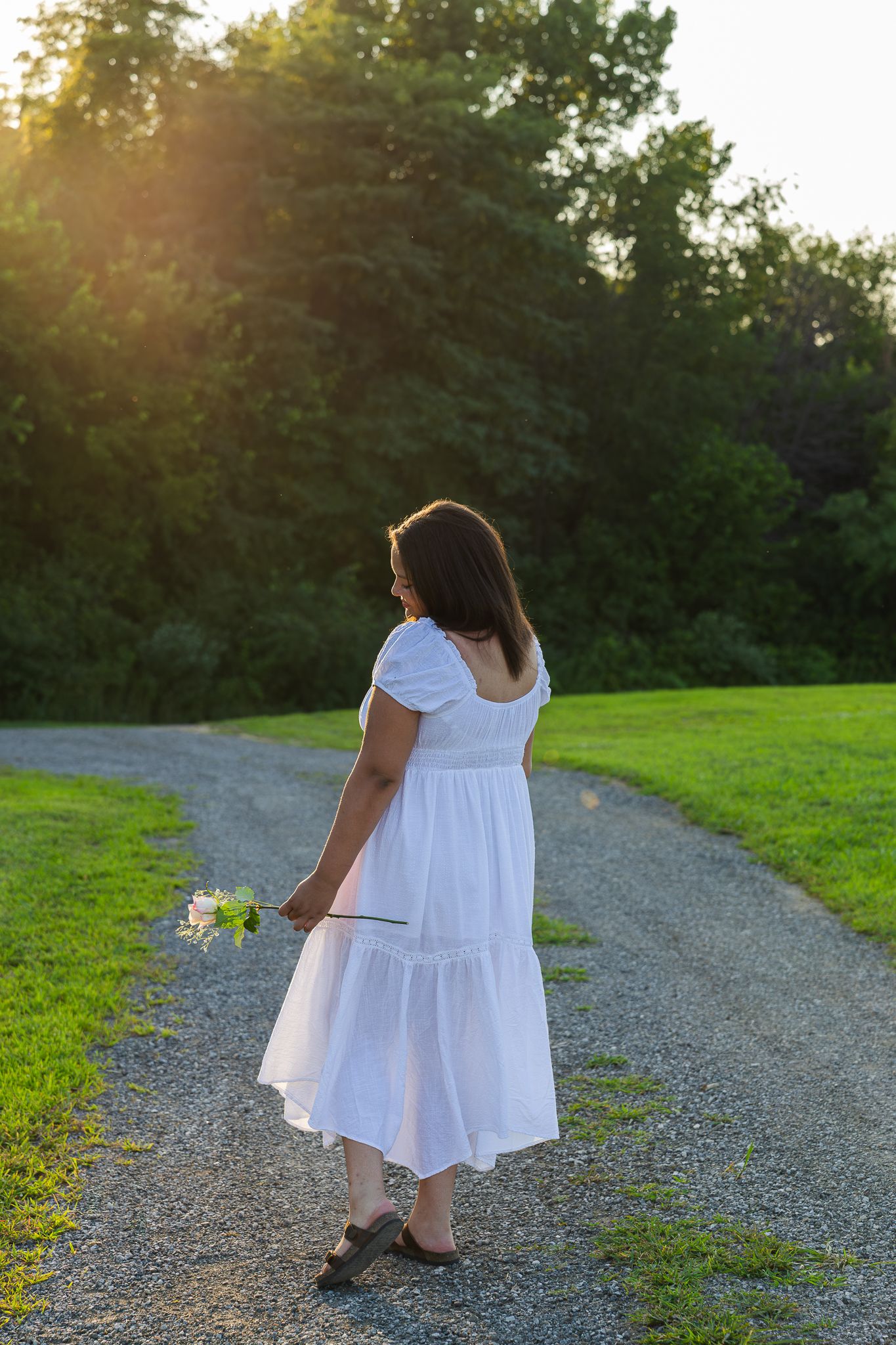 A person in a flowing white dress stands on a gravel path at sunset holding wildflowers amidst green surroundings.