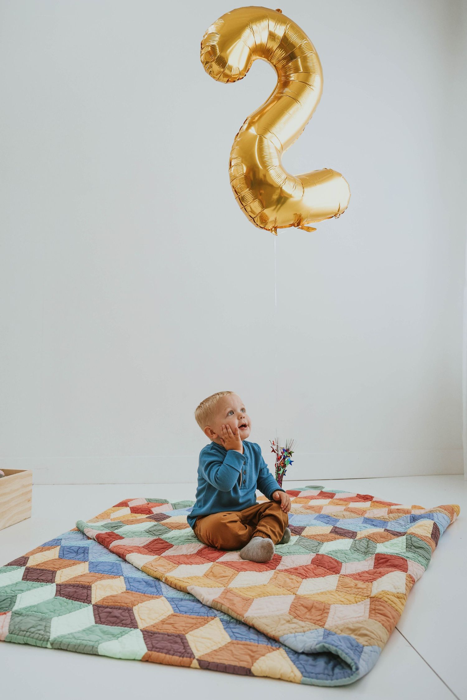 Child sitting on colorful quilt beneath gold number 2 balloon against white background.