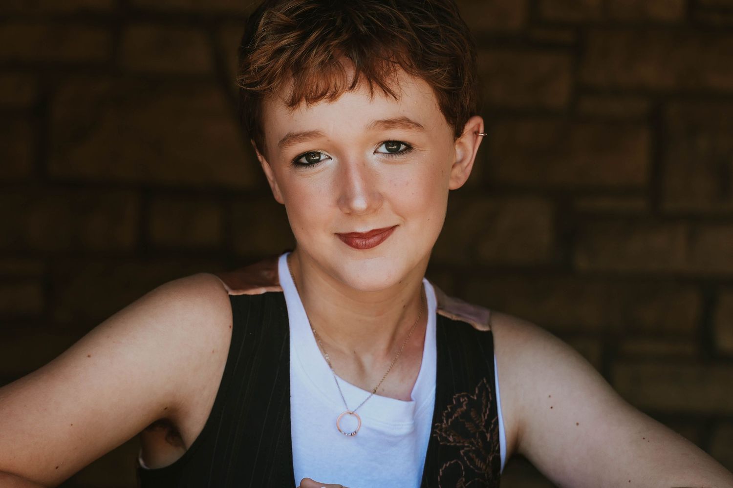 Portrait of a person in white tank top and dark vest against brick wall background.