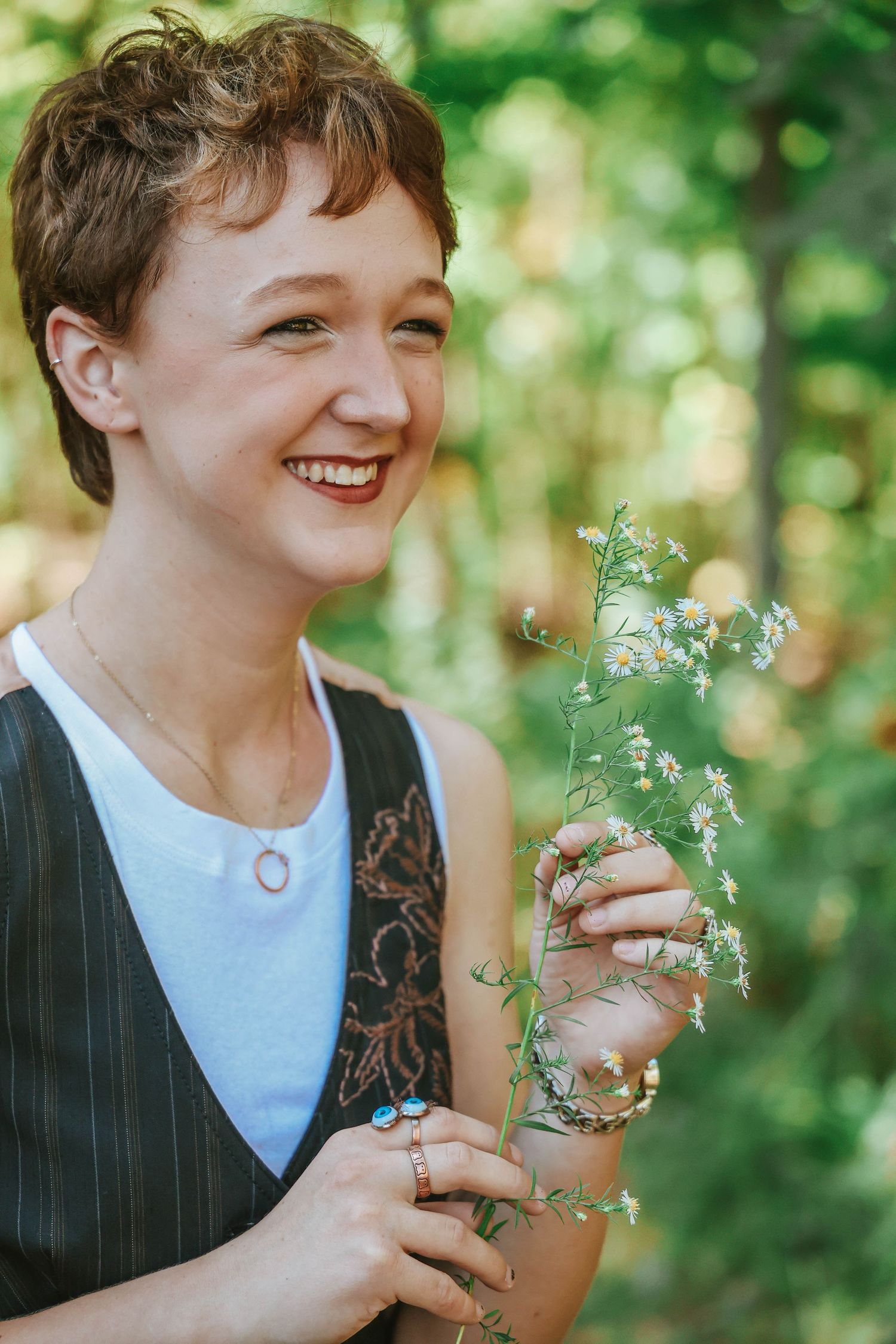 Person in white top and pinstripe vest smiles while holding delicate foliage in sunlit forest.