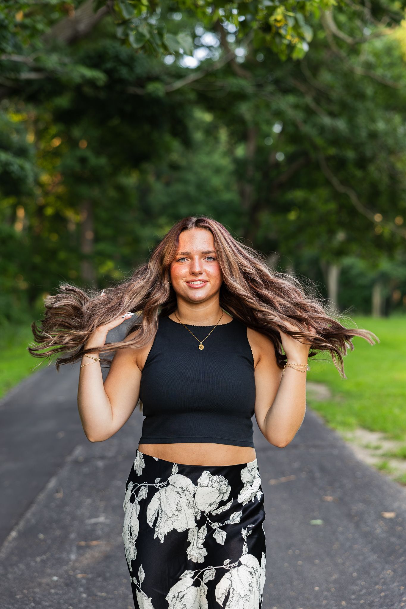 A person in a black crop top and floral pants stands on a tree-lined path while playing with flowing brown hair.