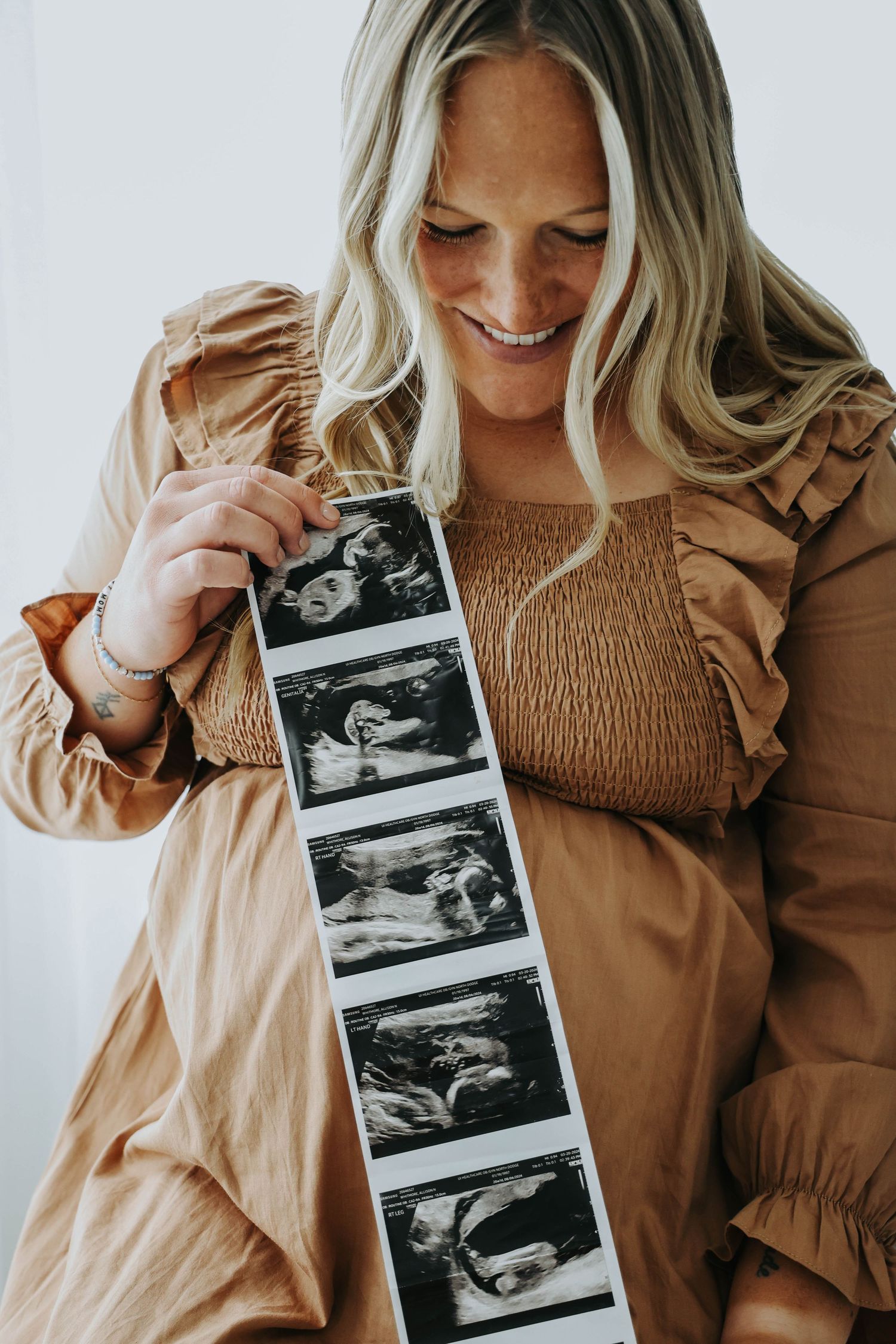 Expectant mother in tan dress holding ultrasound photos while smiling.