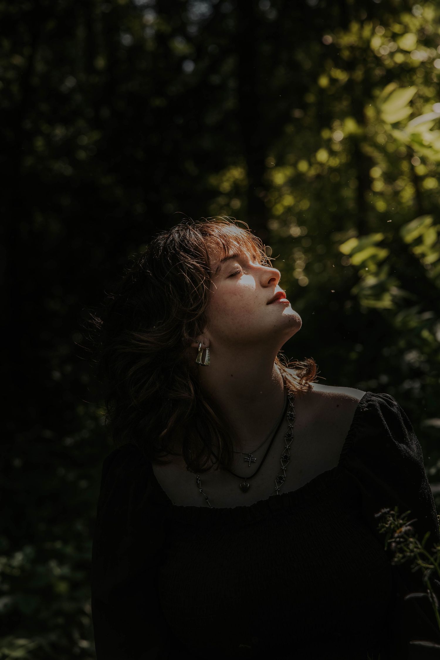 Moody artistic portrait in dark forest with dramatic natural lighting and deep shadows.