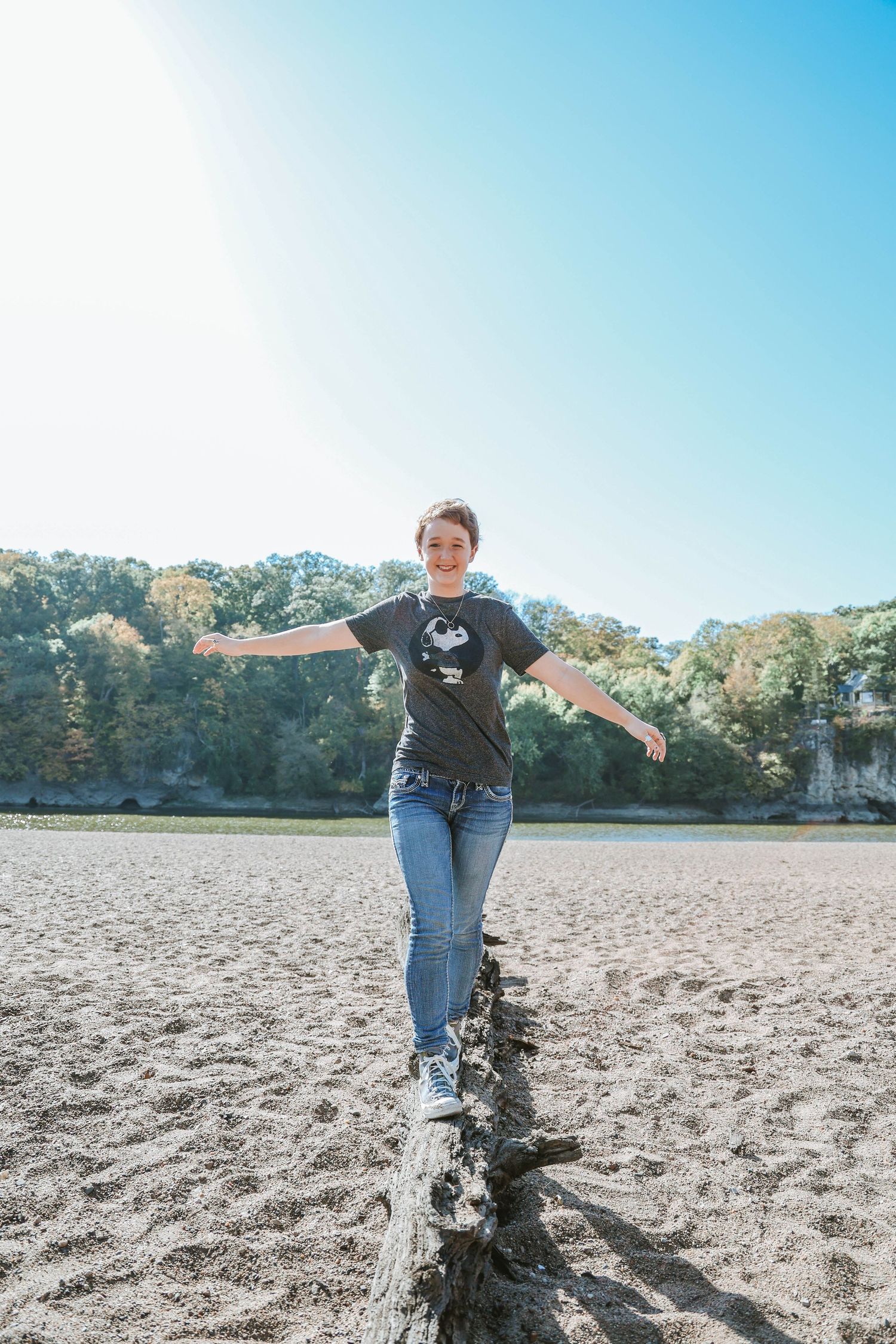Person in casual attire balances on a log at a sandy beach on a sunny day.