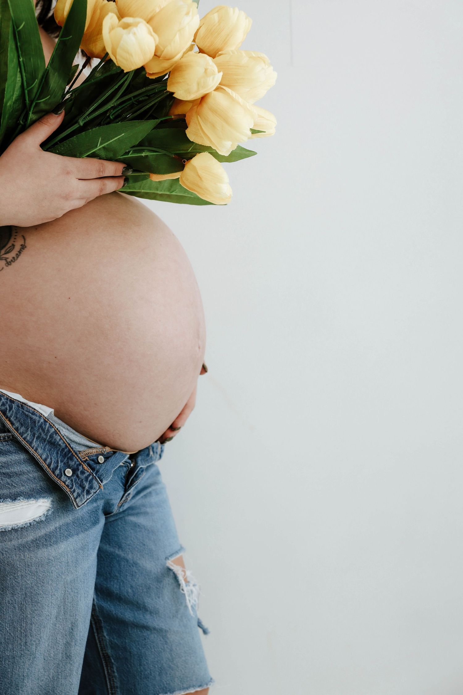 A pregnant belly in ripped jeans holds pale yellow tulips against a white background.