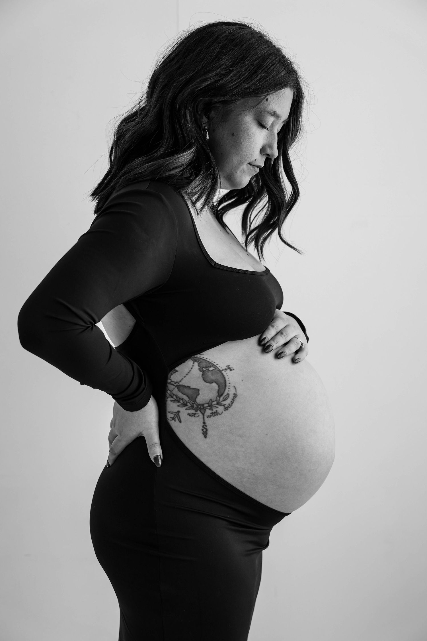 Black and white artistic maternity photo showing a pregnant woman with a tattoo in a black dress on a light background.