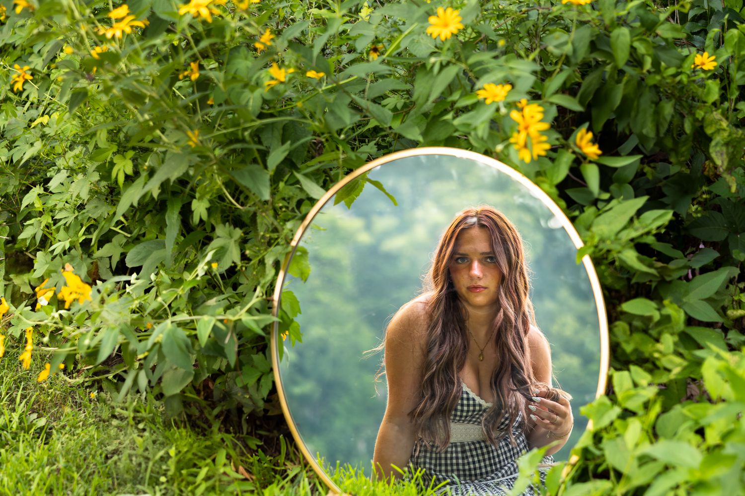 A round mirror reflecting long hair and greenery sits among yellow wildflowers and lush foliage in a garden setting.