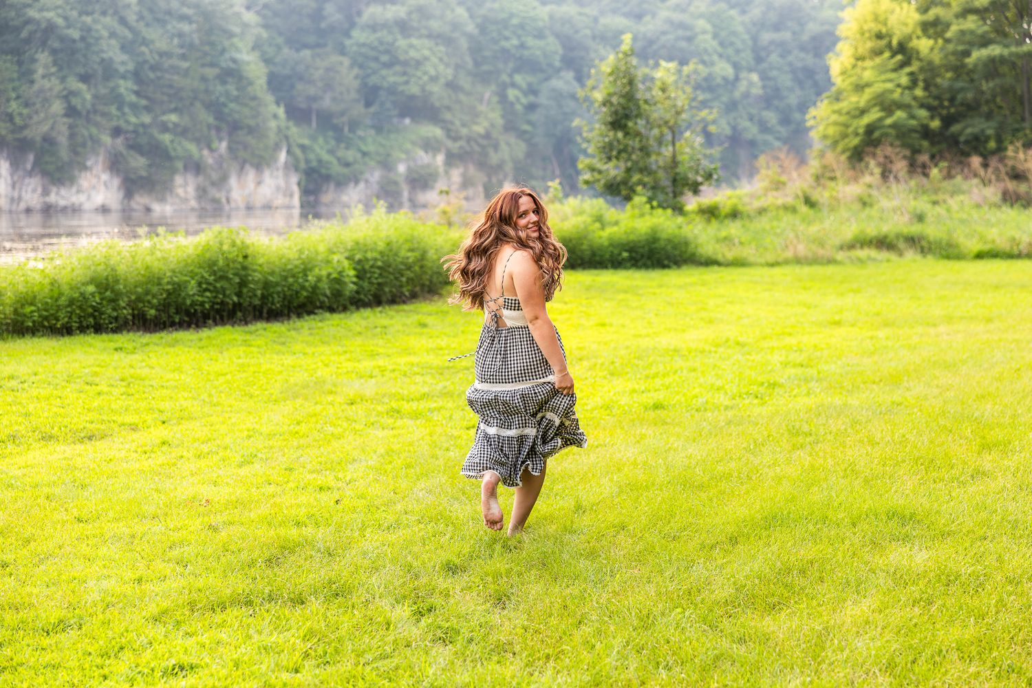 A person twirls in a flowing skirt on a sunny green lawn near a wooded area.
