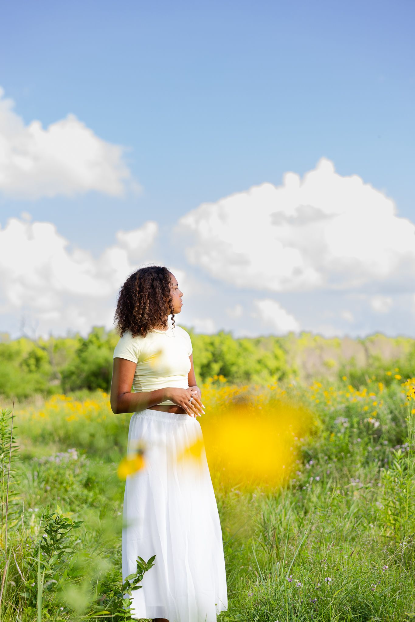 A person in a flowing white dress stands in a sunlit meadow with wildflowers under puffy white clouds.