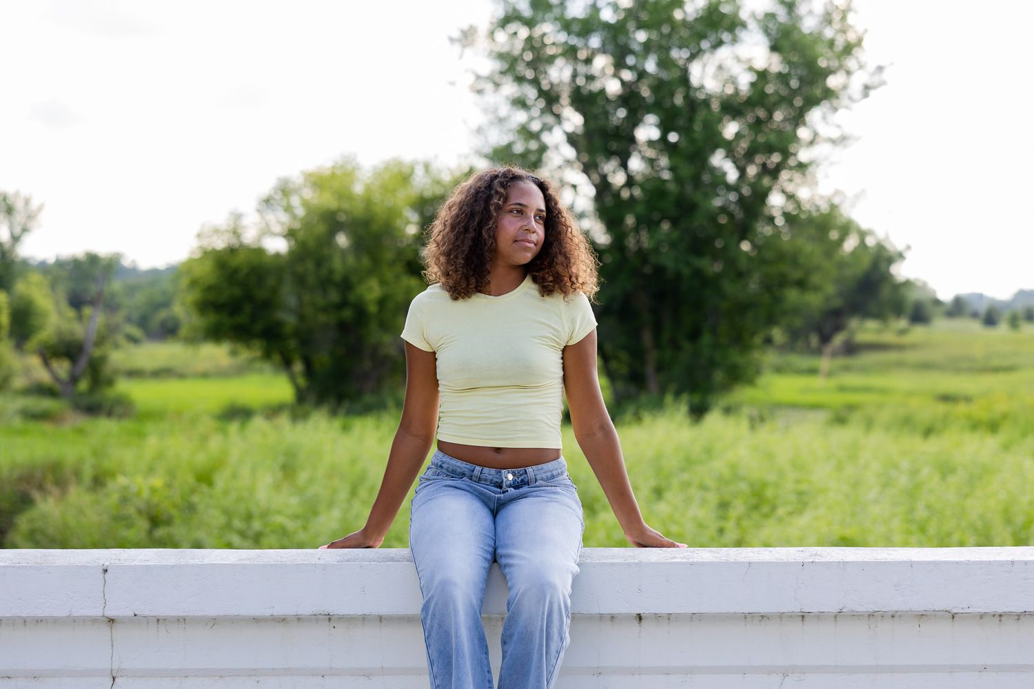 A person wearing light jeans and a white crop top sits on a stone wall in a natural outdoor setting.