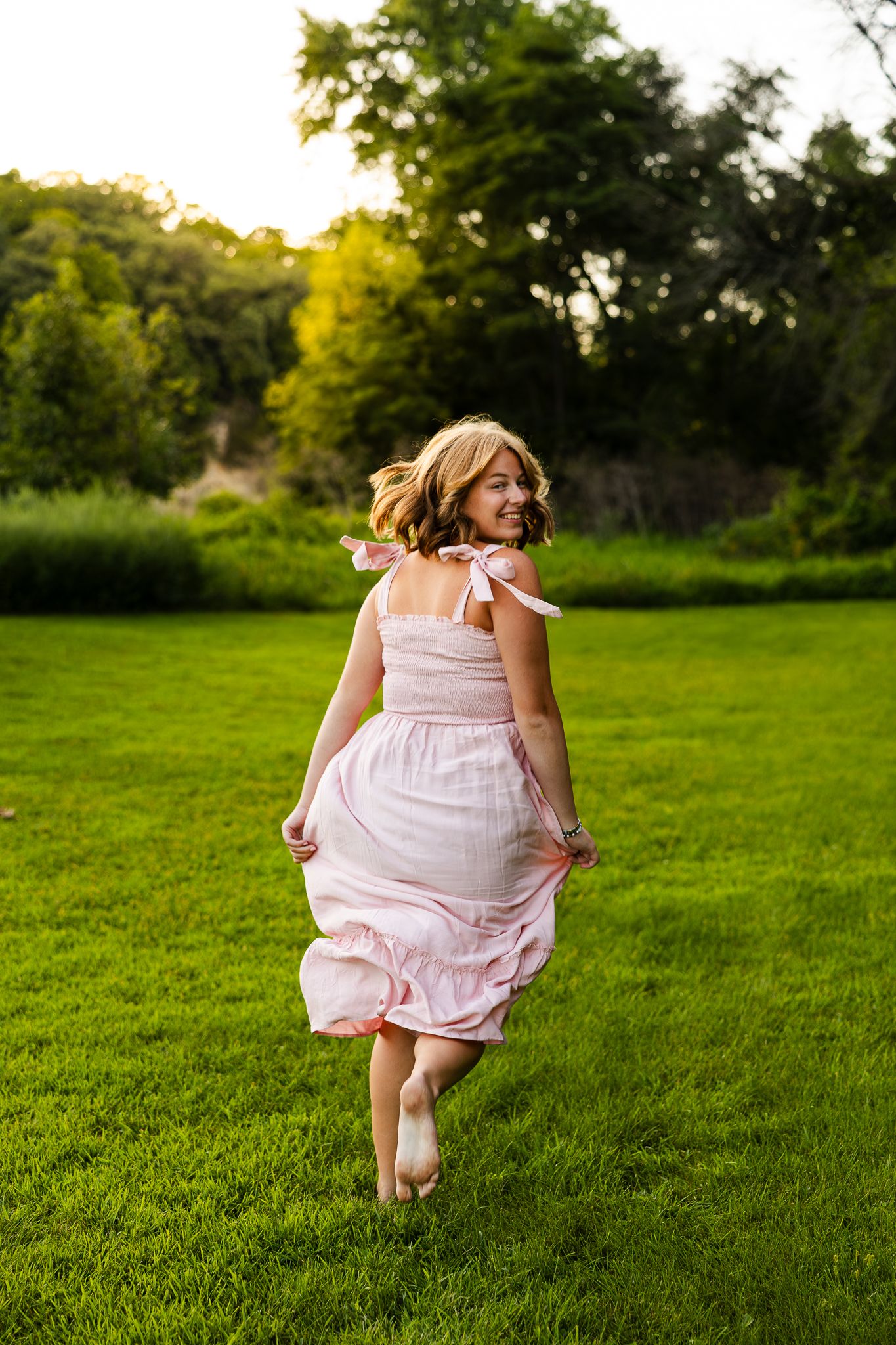 A person in a light pink dress skips joyfully across a lush green lawn on a sunny summer day.