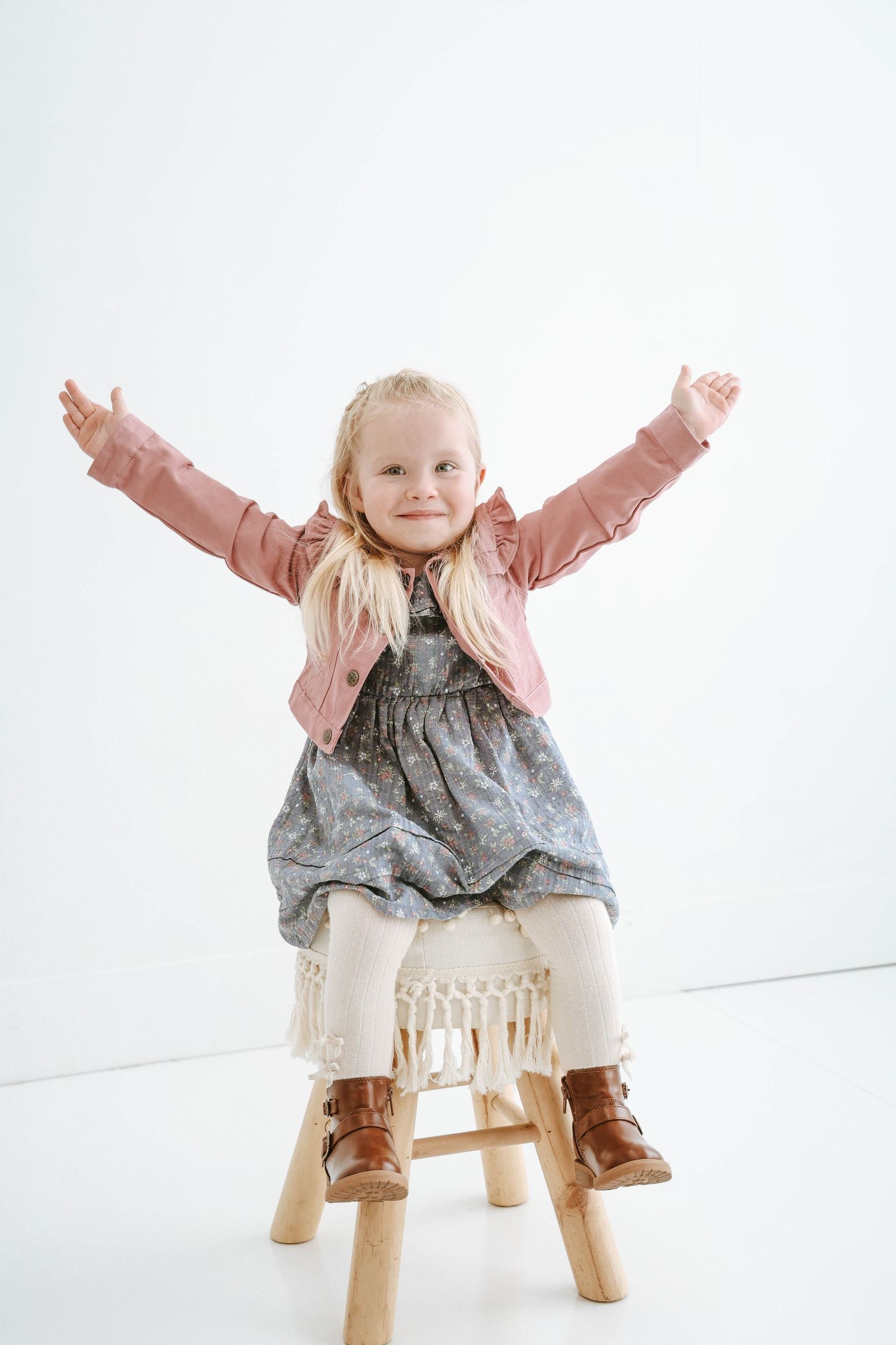 Young child in pink jacket and gray dress sitting triumphantly on wooden stool.
